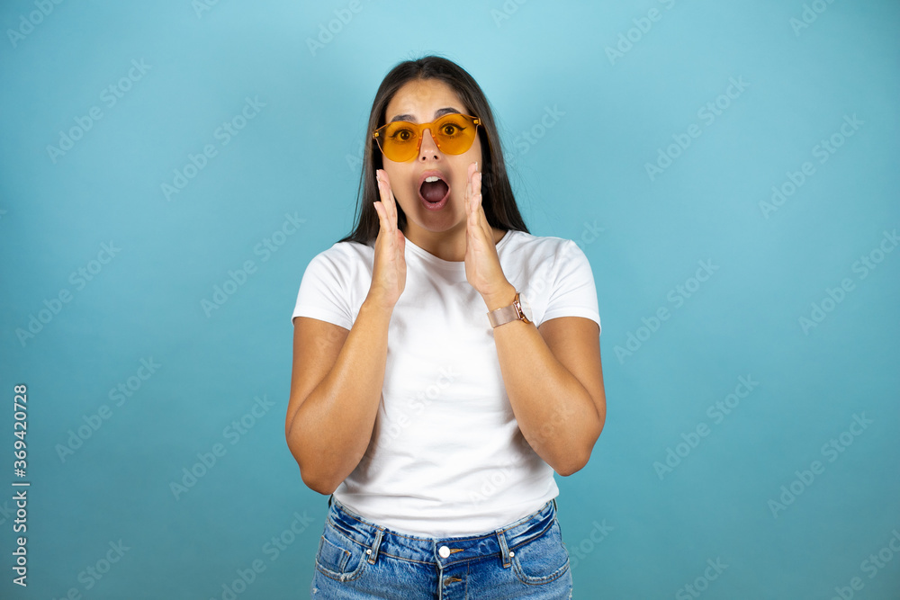 Young beautiful woman wearing sunglasses standing over blue isolated background with her hands over her mouth and surprised Young beautiful woman wearing sunglasses standing over blue isolated background with her hands over her mouth and surprised