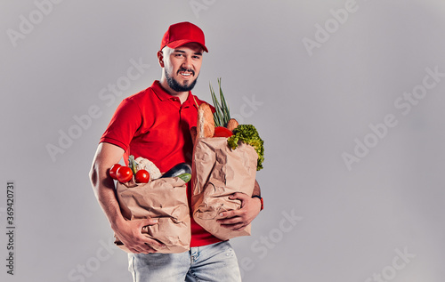 Bearded young man in red T-shirt and cap holding two large heavy bags with food isolated on gray background. Fast food delivery to your home.