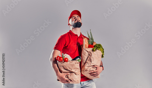 Bearded young man in red T-shirt and cap holding two large heavy bags with food isolated on gray background. Fast food delivery to your home.