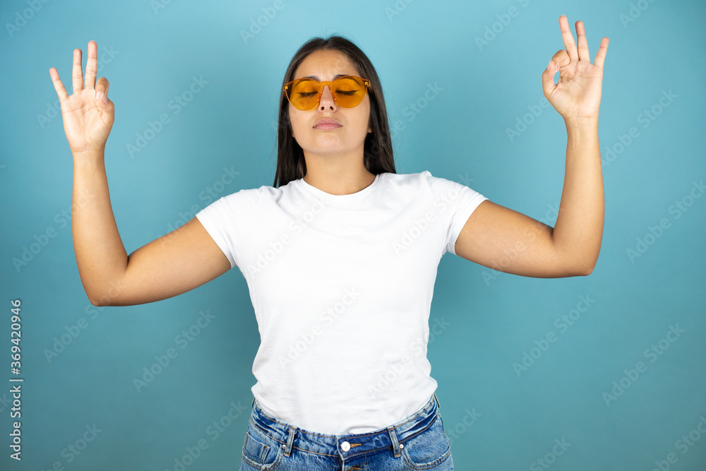 Young beautiful woman wearing sunglasses over isolated blue background relax and smiling with eyes closed doing meditation gesture with fingers Young beautiful woman wearing sunglasses over isolated blue background relax and smiling with eyes closed doing meditation gesture with fingers