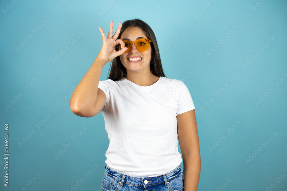 Young beautiful woman wearing sunglasses standing over isolated blue background doing ok gesture shocked with smiling face, eye looking through fingers Young beautiful woman wearing sunglasses standing over isolated blue background doing ok gesture shocked with smiling face, eye looking through fingers