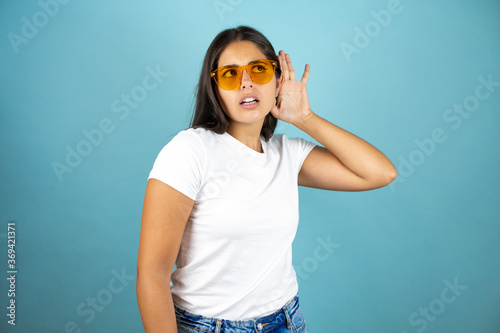 Young beautiful woman wearing sunglasses standing over isolated blue background surprised with hand over ear listening an hearing to rumor or gossip © Irene