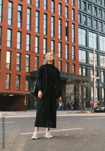 Canvas Print Full length portrait of a fashionable girl outdoors on the background of modern architecture, looking and posing at camera