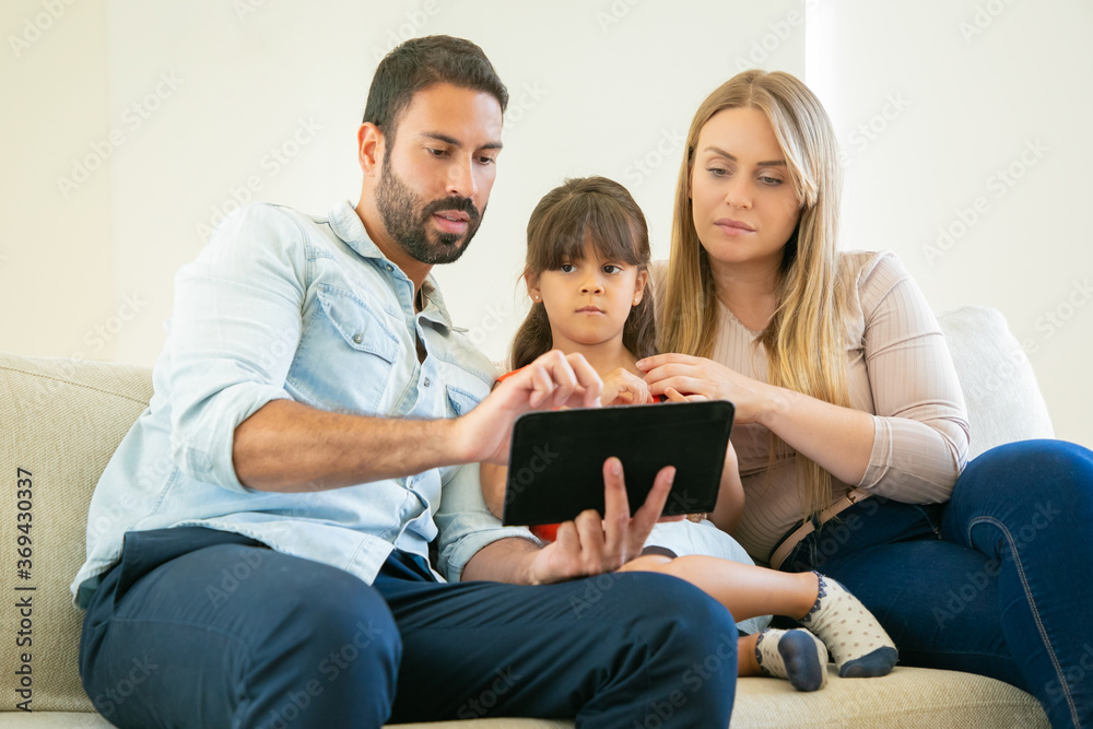 Young couple and cute girl sitting on couch, using online app on tablet, watching video or reading together. Front view. Internet and home entertainment concept Young couple and cute girl sitting on couch, using online app on tablet, watching video or reading together. Front view. Internet and home entertainment concept