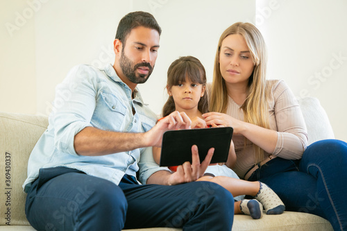 Young couple and cute girl sitting on couch, using online app on tablet, watching video or reading together. Front view. Internet and home entertainment concept