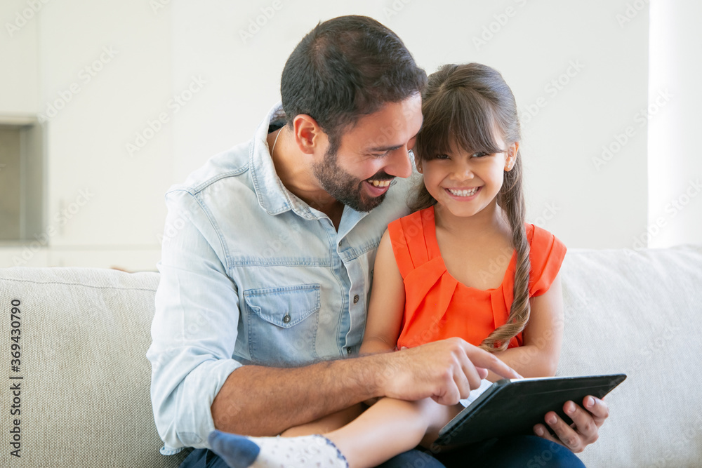 Joyful girl sitting on her dads lap, looking at camera and laughing. Father enjoying time with daughter while using tablet. Internet and technology concept Joyful girl sitting on her dads lap, looking at camera and laughing. Father enjoying time with daughter while using tablet. Internet and technology concept
