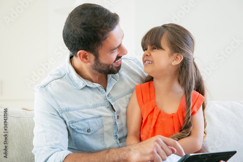 Happy girl sitting on her dads lap and laughing. Father enjoying time with daughter while holding tablet. Family and parenthood concept