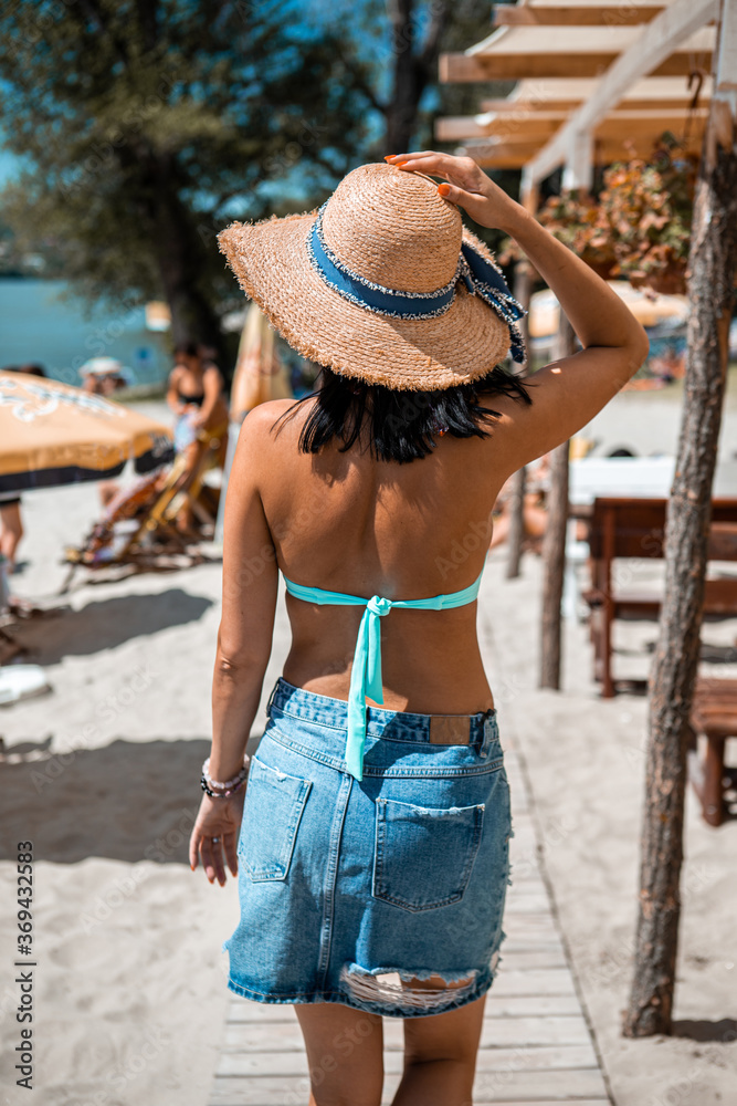 A back view of a tanned young woman walking on the beach. She is wearing a denim skirt and a blue swimsuit, and she is wearing a knitted hat.
