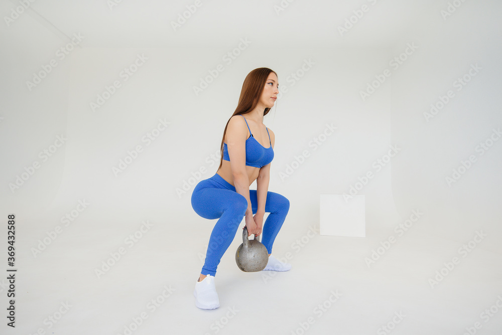 Fototapeta premium Sexy young girl performs exercises with a weight in a blue tracksuit on a white background. Fitness, healthy lifestyle