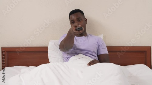 Portrait of African man in bed turns on TV with remote controller. Caucasian woman's legs appear in front of boyfriend and taking off panties. Guy throws remote away and calling girl to him