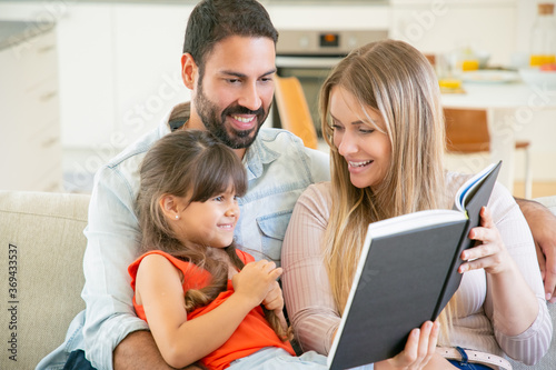 Cheerful parents and little black haired girl sitting on couch in living room, reading book together and laughing. Childhood or literature concept