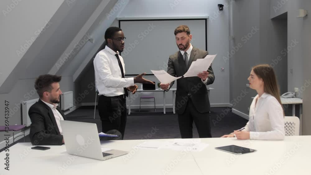 Angry boss scolding dark skinned employee for bad work result, male director yelling at incompetent subordinate blaming for mistake in financial document team meeting.