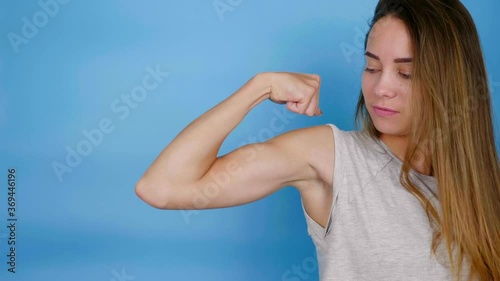 Beautiful strong powerful athletic smiling woman showing biceps muscles on hand, thumb up, like, kiss. Sports fitness girl in gray t-shirt on blue background with copy space. 4K slow motion footage photo