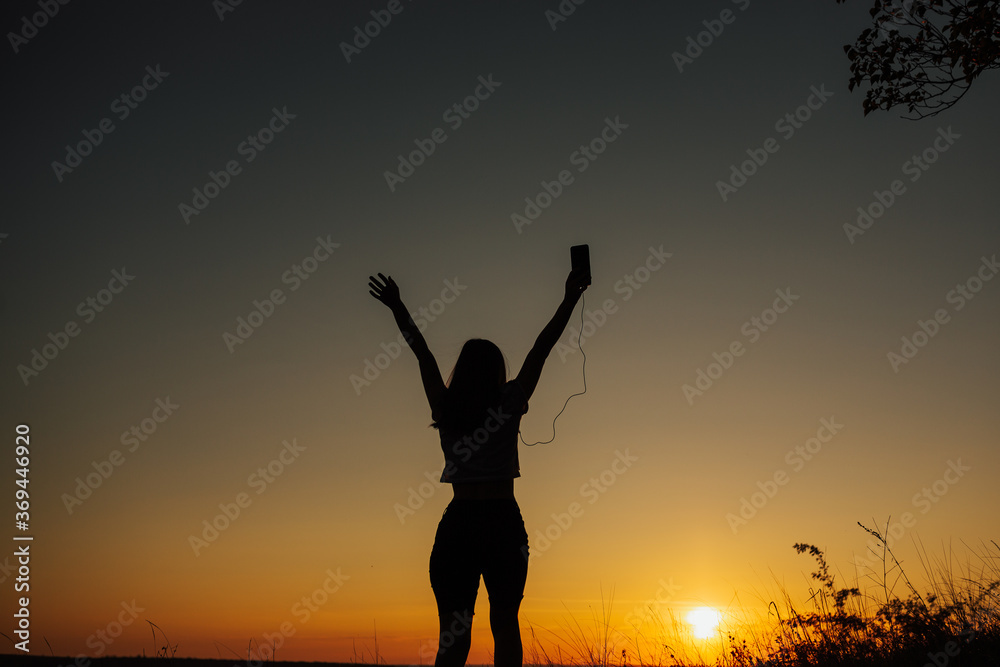 Free girl in headphones listen to music and dancing with hands up at beautiful sunset in the field.  Excited girl dancing and listening music with headphones and smart phone. Copy space.