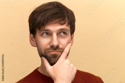 Hard choice. Photo of young man with beard wearing sweater, holds chin, purses lips with clueless expressions, doubts about something, posing against yellow background photo