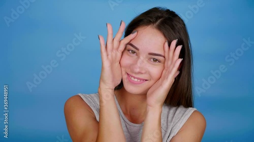 Beautiful positive woman hiding, covering eyes, face with palms, hands. Girl in gray t-shirt opens face and smiles on blue isolated background with copy space. 4K slow motion footage