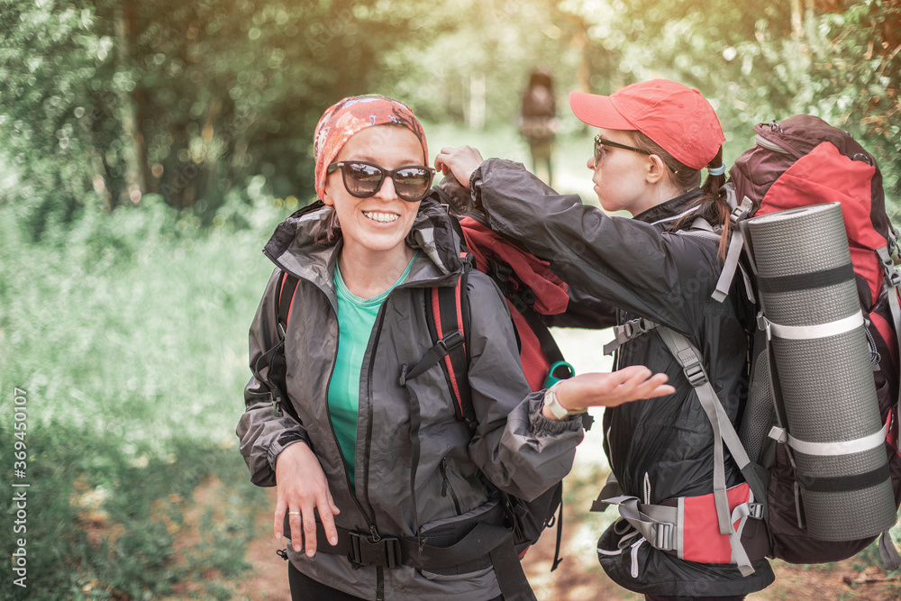 Two female tourists travel through the forest with trekking backpacks. Friends check each other's equipment and have fun