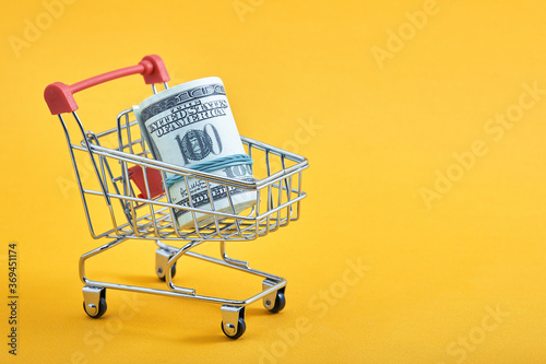 Mini shopping cart with 100 dollar banknotes inside on yellow background. Trolley and money. Financial crisis or shopaholic concept. Flat lay, copy space.