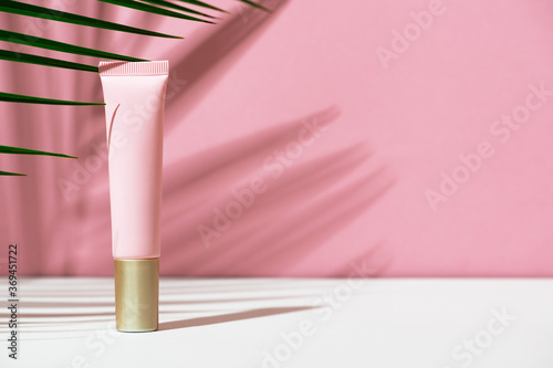 Foundation in a pink plastic tube on a white, pink background with leaves and shadow of a tropical palm tree. Women's cosmetic accessory for make-up, tint, fluid. Mockup, identity, copy space.