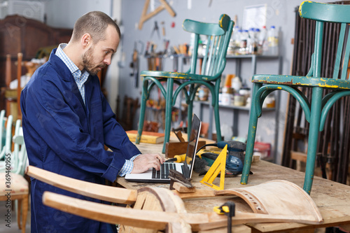 Professional young man carpenter using laptop while repairing antique furniture in workshop © JackF