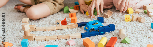 partial view of nanny and child near multicolored cubes and toy car on floor, panoramic shot