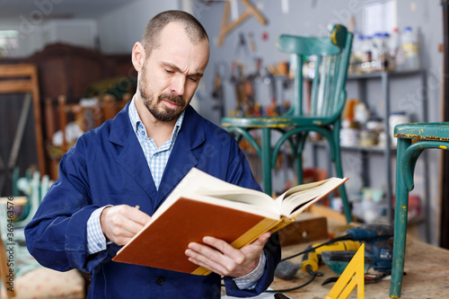 Fotografi Portrait of positive professional man carpenter standing with book in furniture repair shop