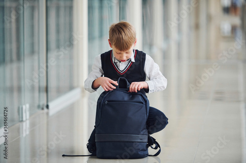 Little school boy in uniform sitting in corridor with backpack