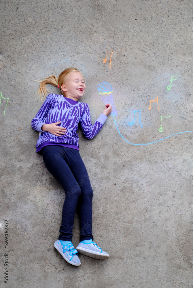 Fototapeta premium Happy little girl have fun introducing herself to the singer profession. The concept of dreaming about a future profession. The child lies on the ground, top view. Childhood and dreams concept.
