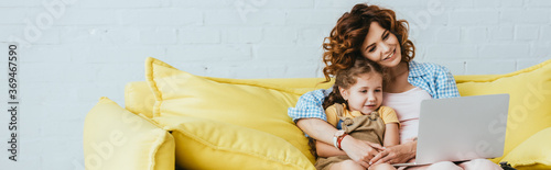 horizontal image of smiling babysitter and child sitting on sofa and using laptop