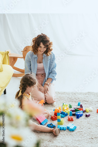 selective focus of smiling babysitter looking at cute kid playing with building blocks on floor