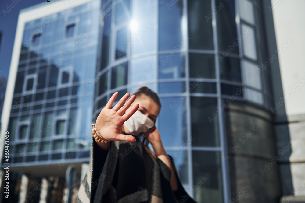 Young woman in protective mask standing outdoors near business building at sunny daytime. Conception of coronavirus