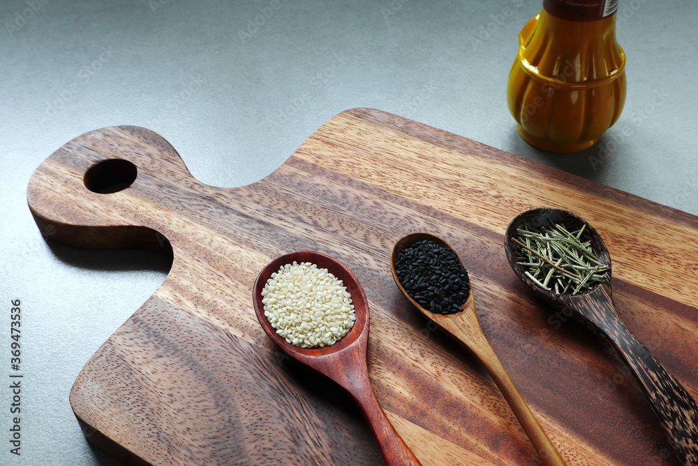 The white sesame, black sesame and rosemary in the wooden spoon on the wooden tray with a bottle of turmeric powder and garlic on the gray table