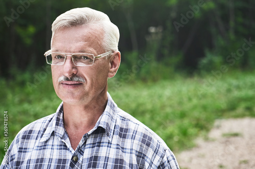 Close-up portrait of handsome gray-haired adult man with mustache and glasses smiling on the background of a summer forest park