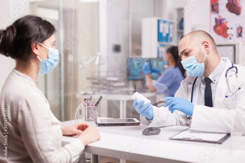 Doctor with face mask against covid-19 holding pills for patient during consultation in hospital office. Medic wearing white coat and stethoscope.