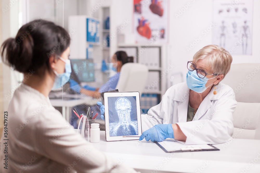 Obraz premium Lekarz z maską agasint covid-19, wskazujący na tablet pc z prześwietleniem mózgu podczas konsultacji z pacjentem.