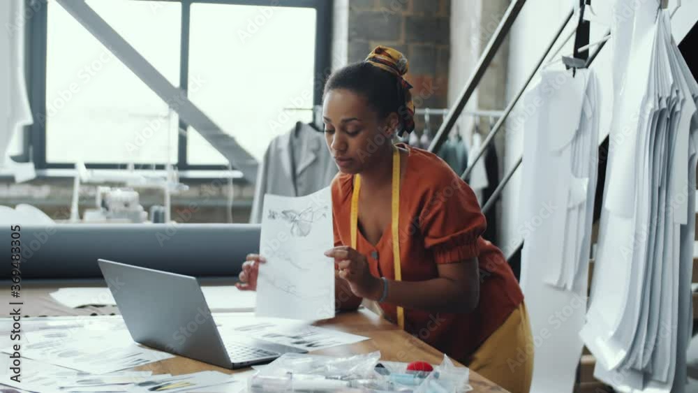 Arc shot of African American female designer smiling and discussing fashion sketches on video call on laptop while working on new collection in atelier