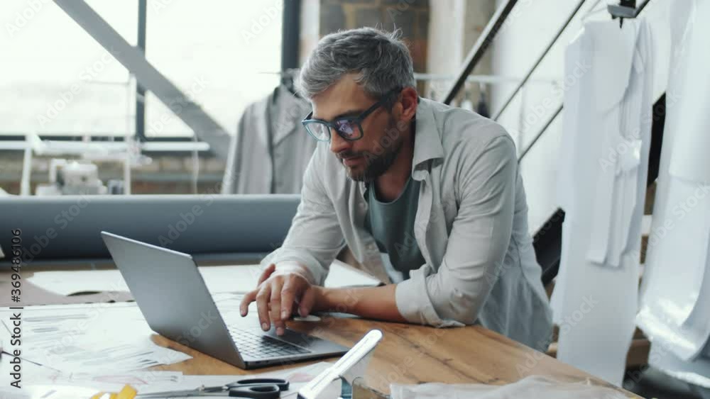 Arc shot of handsome bearded tailor leaning on table, using laptop and then looking at camera with confidence while posing in sewing studio Arc shot of handsome bearded tailor leaning on table, using laptop and then looking at camera with confidence while posing in sewing studio