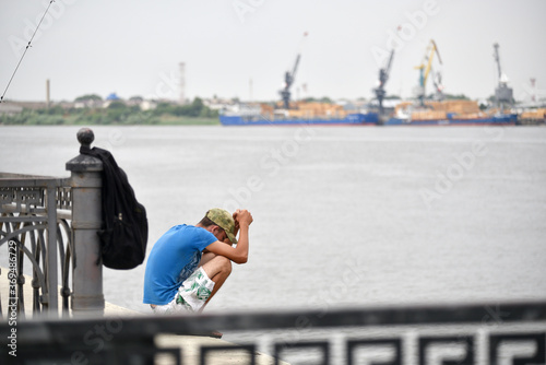 A young guy in a blue T-shirt is sad on the river bank against the background of the river port. Concept for staff reduction, layoff, crisis photo