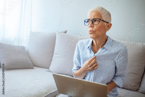 Low angle of restless mature woman holding laptop and having hot flash. Shortness of breath. Unhappy mature woman sweating and touching shirt. During menopause.