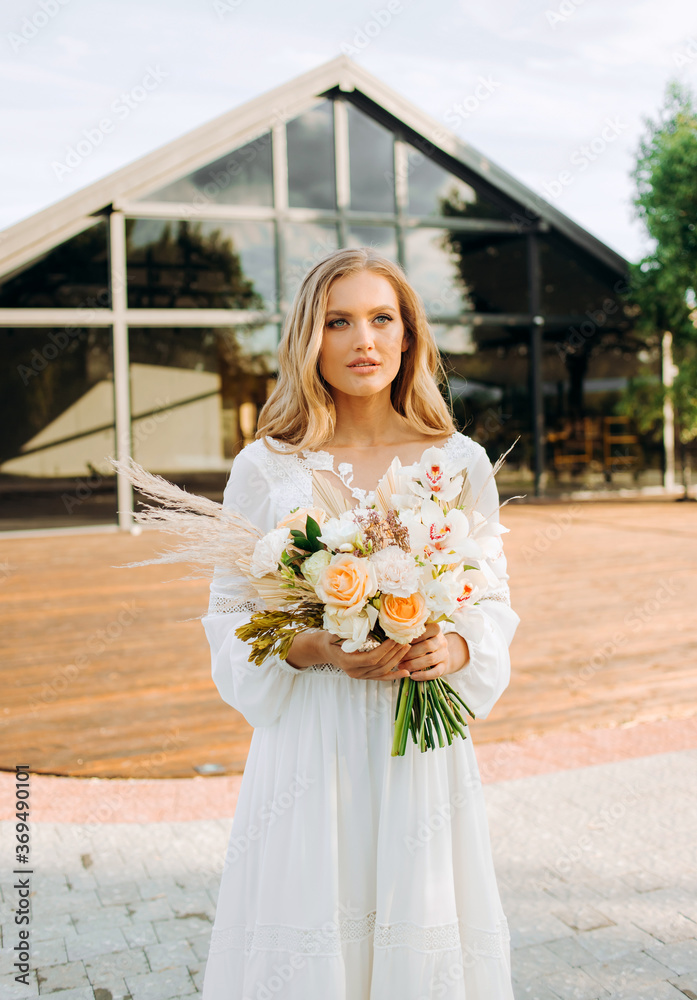 Fototapeta premium Beautiful blonde bride holding a bouquet in her hands on the background of a modern building