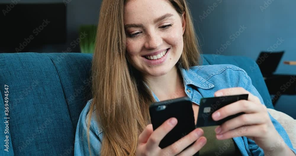 Close Up of Beautiful young Woman with Freckles is shopping online with Credit Card and Smartphone. Young Woman is Shopping from Home, getting Positive Emotions while using her Smartphone. Indoor. Close Up of Beautiful young Woman with Freckles is shopping online with Credit Card and Smartphone. Young Woman is Shopping from Home, getting Positive Emotions while using her Smartphone. Indoor.