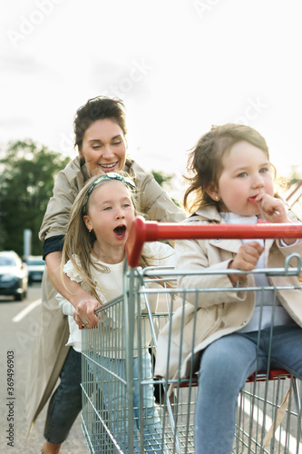 Happy mother and her daughters are having fun with a shopping cart on a parking lot beside a supermarket.