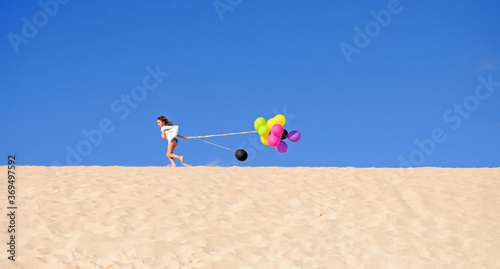 a little girl in a white dress holds colorful balloons and runs on sand dunes of Corralejo, Fuerteventura island