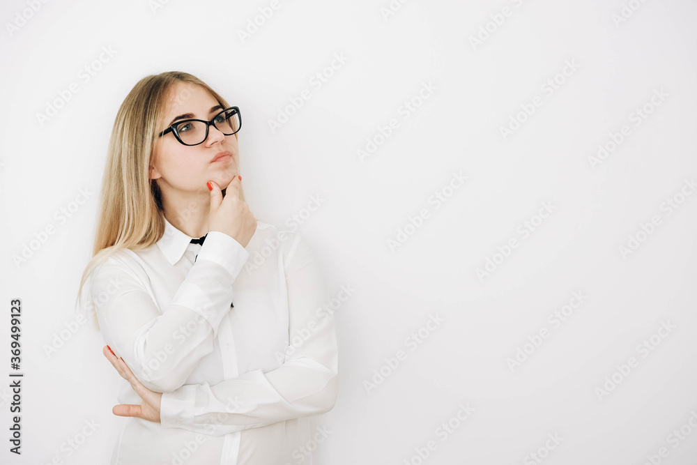 Beautiful blonde girl wears glasses stylish white shirt with a pensive face on a white wall background. Female has clever expression, keeps fore finger near mouth, tries to remember material she