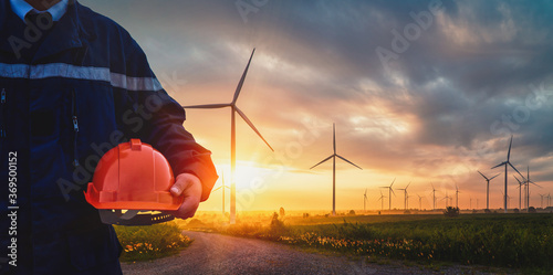 Technician Engineer in uniform and holding orange safety helmet with standing and checking wind turbine power farm Power Generator Station. Clean energy and environment 