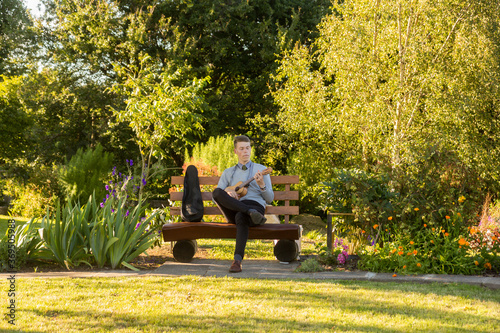 Photos Young man sitting on bench in the park and playing ukulele.