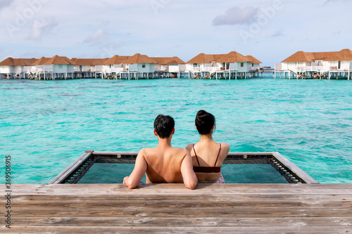 Back of view of romantic couple in love sitting on pier together with beautiful sea. Elegant honeymoon traveler couple hugging on a wooden jetty and enjoy their tropical holiday enjoying ocean view. © Avirut S.