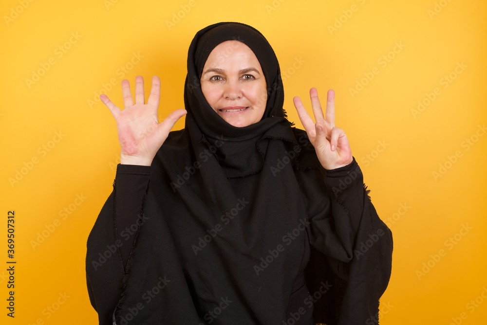 Beautiful middle aged muslim woman wearing black hijab over yellow background showing and pointing up with fingers number eight while smiling confident and happy. Beautiful middle aged muslim woman wearing black hijab over yellow background showing and pointing up with fingers number eight while smiling confident and happy.
