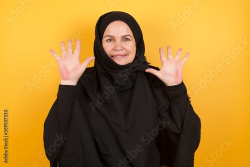 Beautiful middle aged muslim woman wearing black hijab over yellow background showing and pointing up with fingers number ten while smiling confident and happy.