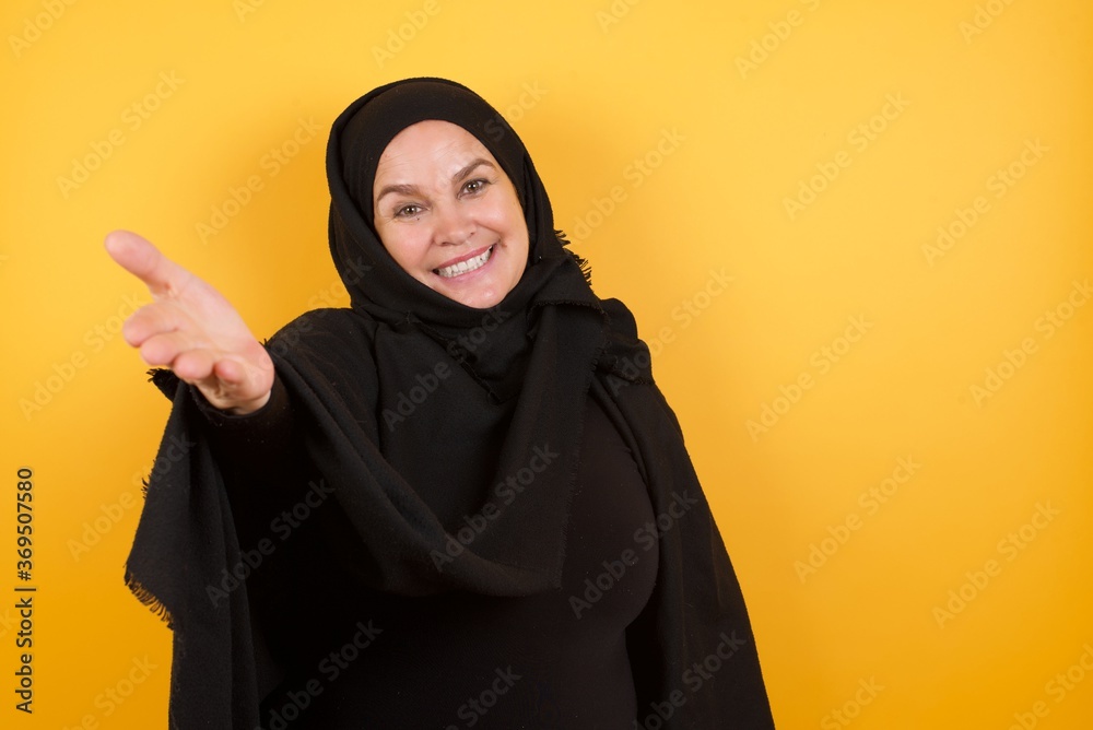 Beautiful middle aged muslim woman wearing black hijab over yellow background smiling friendly offering handshake as greeting and welcoming. Successful business.